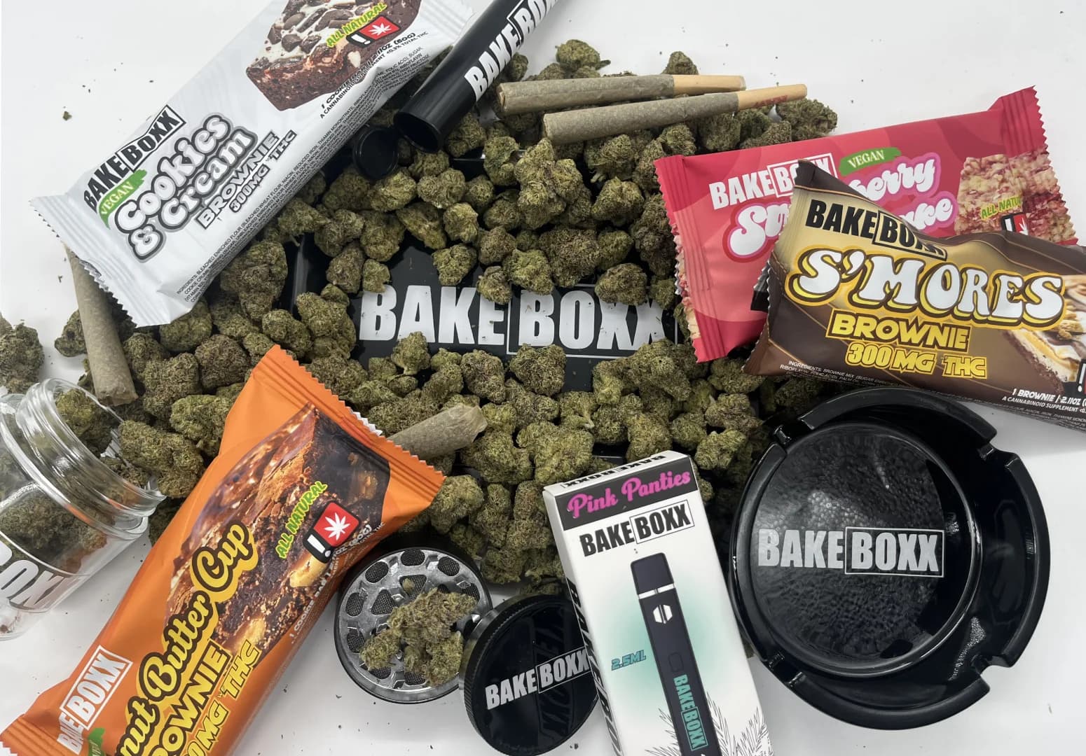 BakeBoxx. Collection of buds, prerolls, and edibles stacked up one on top of anotherm featuring Strawberry Shortcake, Peanut Butter Cup Brownie, Cookies & Cream Brownie, and S'Mores Brownie.